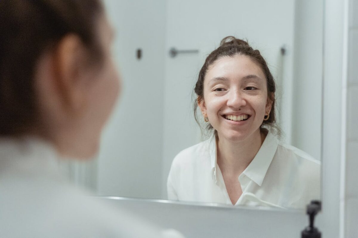 A woman in a white shirt smiles warmly at her reflection in a bathroom mirror.