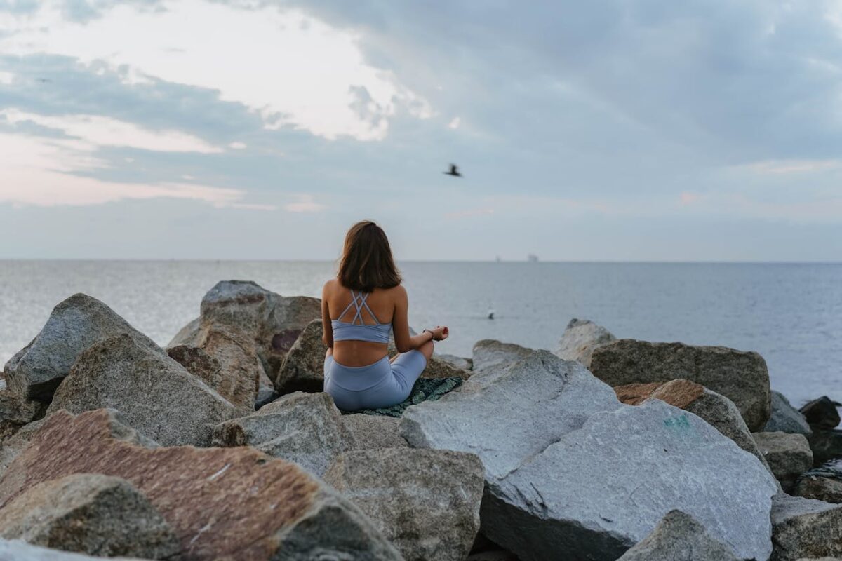 A woman meditates on rocky beach during sunrise, promoting relaxation and mindfulness.